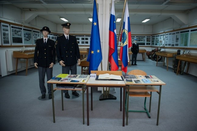 Razstava v Muzeju slovenske policije; miza z zloženskami, v ozadju slovenska, evropska zastava in silhueta policista