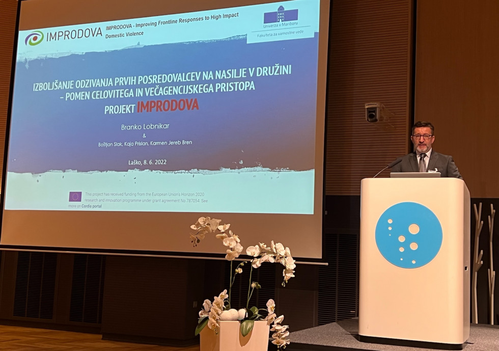 Državni sekretar Branko Lobnikar stoji za belo govornico, na njegovi desni ekran z vsebino predavanja