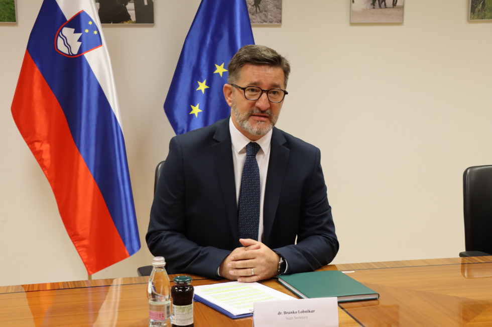 Državni sekretar dr. Branko Lobnikar sedi za mizo, za njim sta slovenska in evropska zastava