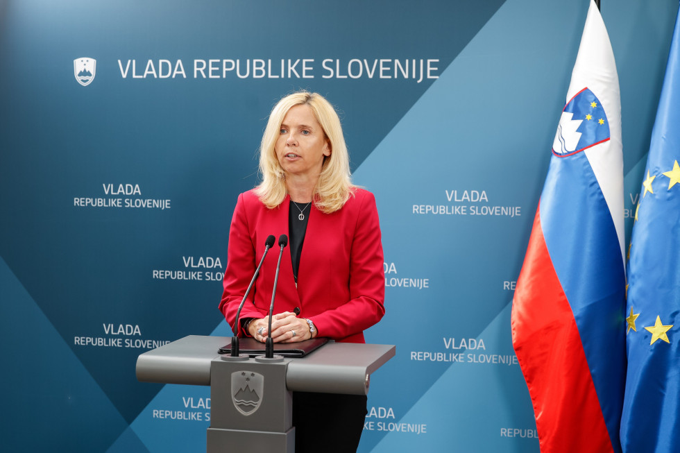 Ministrica Tatjana Bobnar stoji pred modrim ozadjem in predstavlja podrobnosti glede načrta za odstranjevanje žice, pred njo govornica