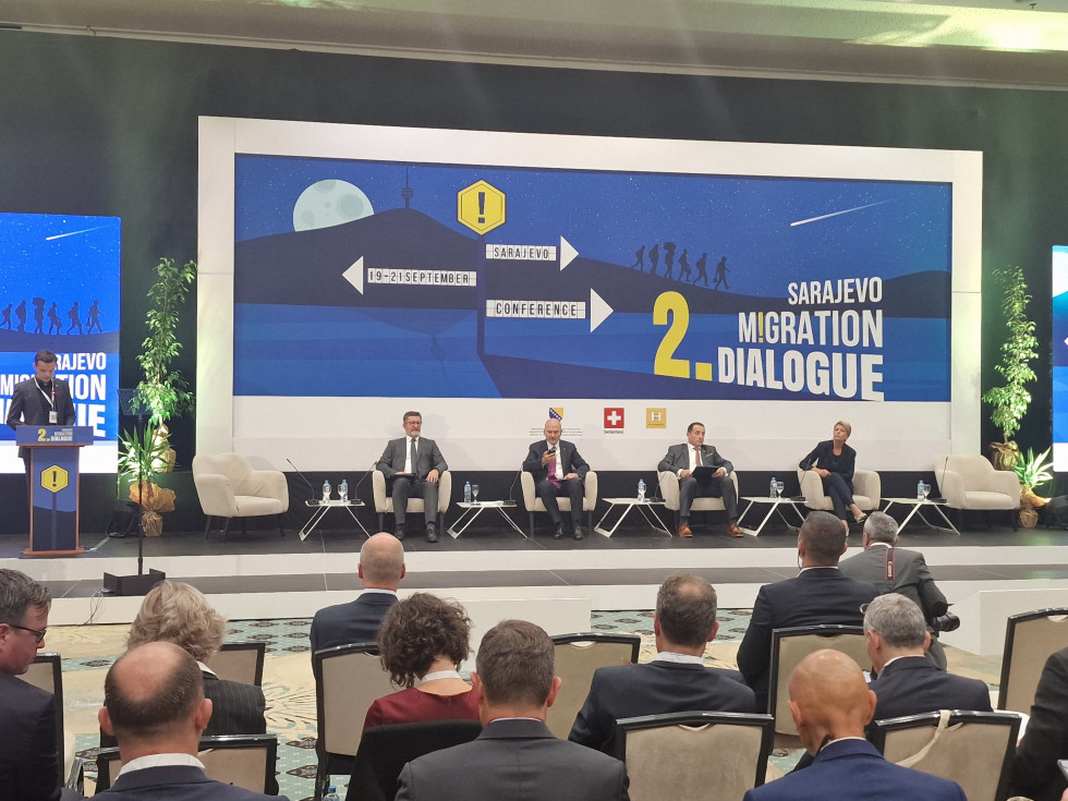 Državni sekretar Branko Lobnikar sedi na stolu na odru s še tremi govorci, za njimi platno z logotipom konference, pred njimi sedijo slušatelji