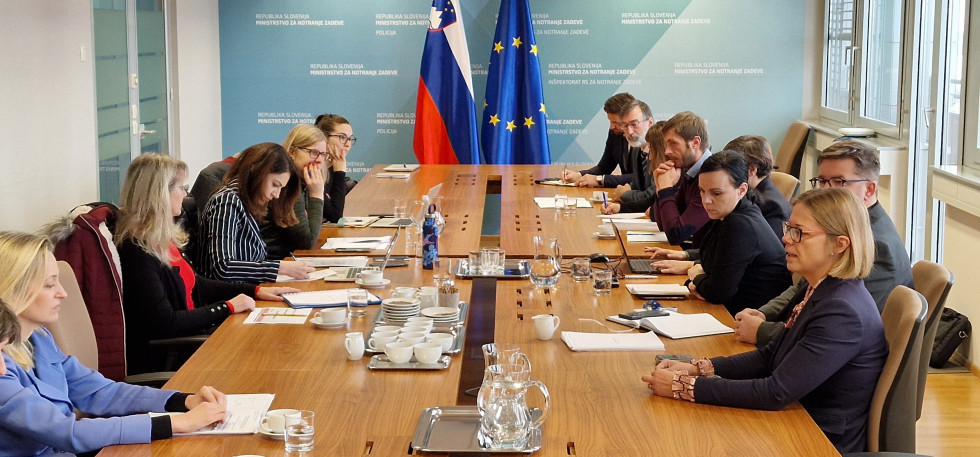 Participants sitting at a large table and listening, with the Slovenian and European flags in the background