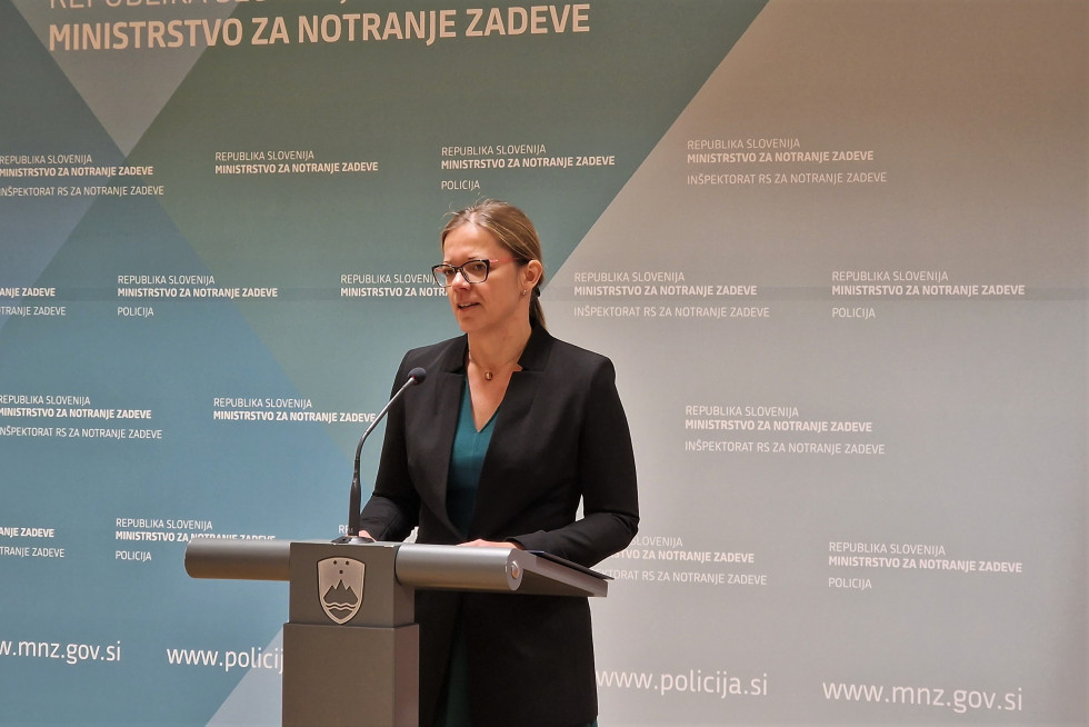 Ministrica Sanja Ajanović Hovnik na novinarski konferenci. Stoji za govornico pred modrim panojem z napisom Ministrstvo za notranje zadeve.