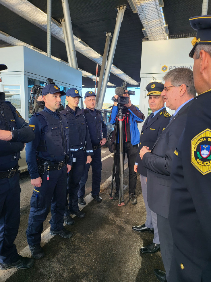 Dogajanje na mejnem prehodu. Tla so asfaltna, na desni strani slike stojijo predstavnik policije in minister Boštjan Poklukar, na levi strani slike stojijo policistka in policisti, ki opravljajo nadzor na mejnem prehodu Obrežje.