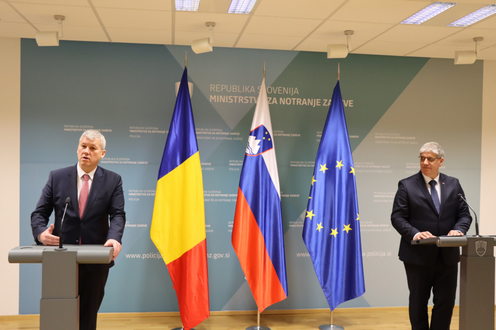Press release by the Ministers of the Interior of Romania and Slovenia. They are standing in the hall, each behind his own grey lectern, behind a blue panel and flags.