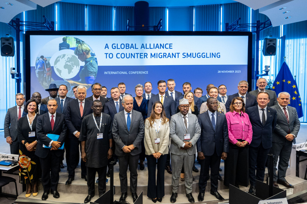 Group photo of participants at the International Conference on the Global Alliance to Combat Migrant Smuggling. They are standing in front of a large blue screen with captions. They are mostly men, about 30 in all.
