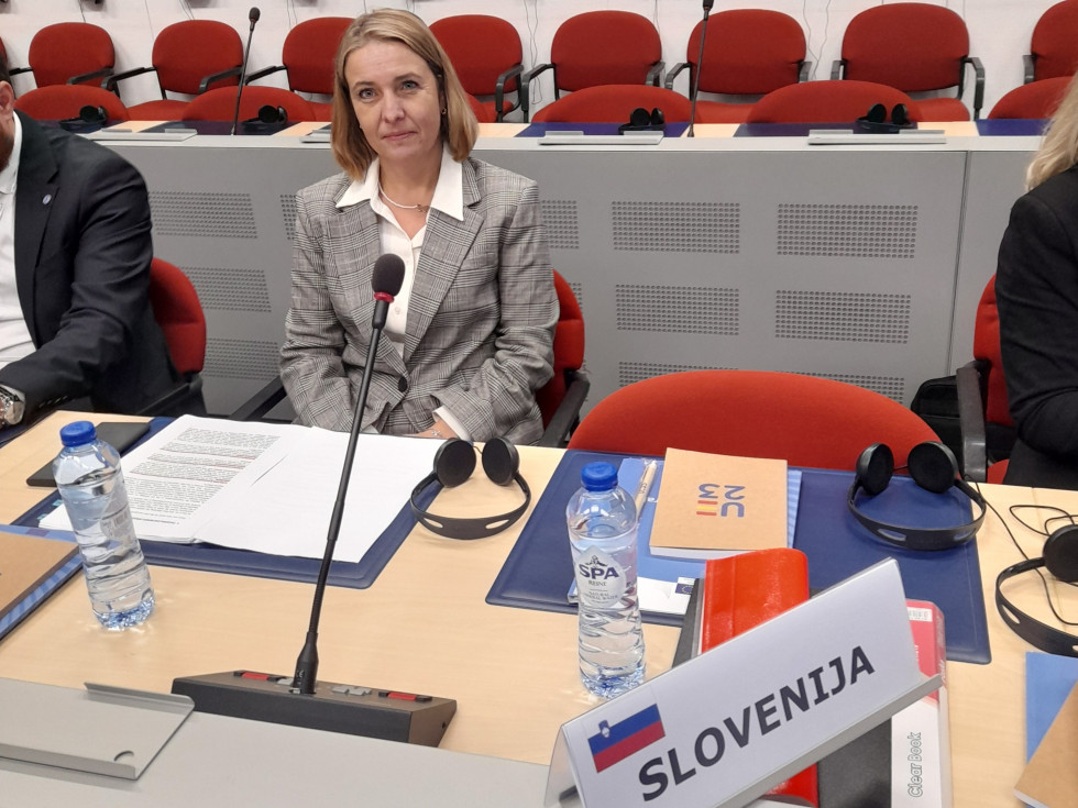 Državna sekretarka Helga Dobrin sedi za svetlo rjavo mizo. Na mizi napis Slovenija, za njo rdeči stoli.