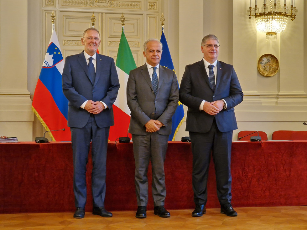 Trije ministri stojijo pred podolgovato mizo, na kateri je rdeč prt. Za njimi zastave Slovenije, Hrvaške, Italije in Evropske unije. Tla so svetlo rjave barve.