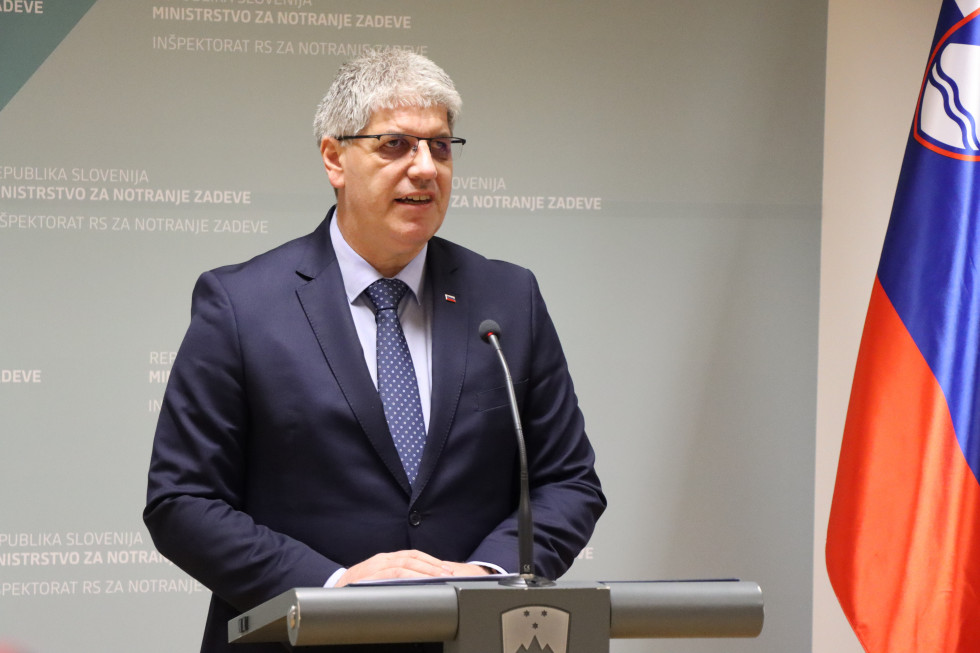 Minister Boštjan Poklukar stands behind a grey lectern, behind him is a blue wall with the Ministry's logos, and in the right-hand corner is a part of the Slovenian flag.