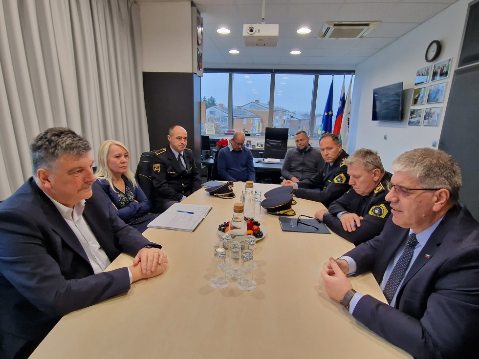 Za dolgo leseno mizo sedijo predstavniki občine, ministrstva in policije, za njimi je okno, v kotu sta postavljeni slovenska zastava in zastava EU..