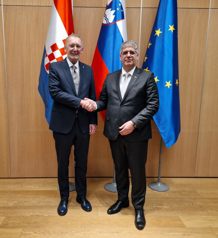 Ministra Boštjan Poklukar in Davor Božinović stojita in se rokujeta pred zastavami Hrvaške, Slovenije in Evropske unije. Prostor je v svetlih tonih.
