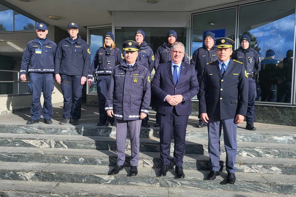 Minister, generalni direktor policije in policisti stojijo zunaj na stopnicah. Sončno vreme.