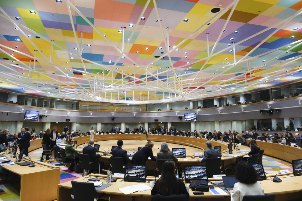 The large meeting room where the Council of the EU meets. People sit at a round table with computers and an empty space in the middle, with a colourful ceiling above them.