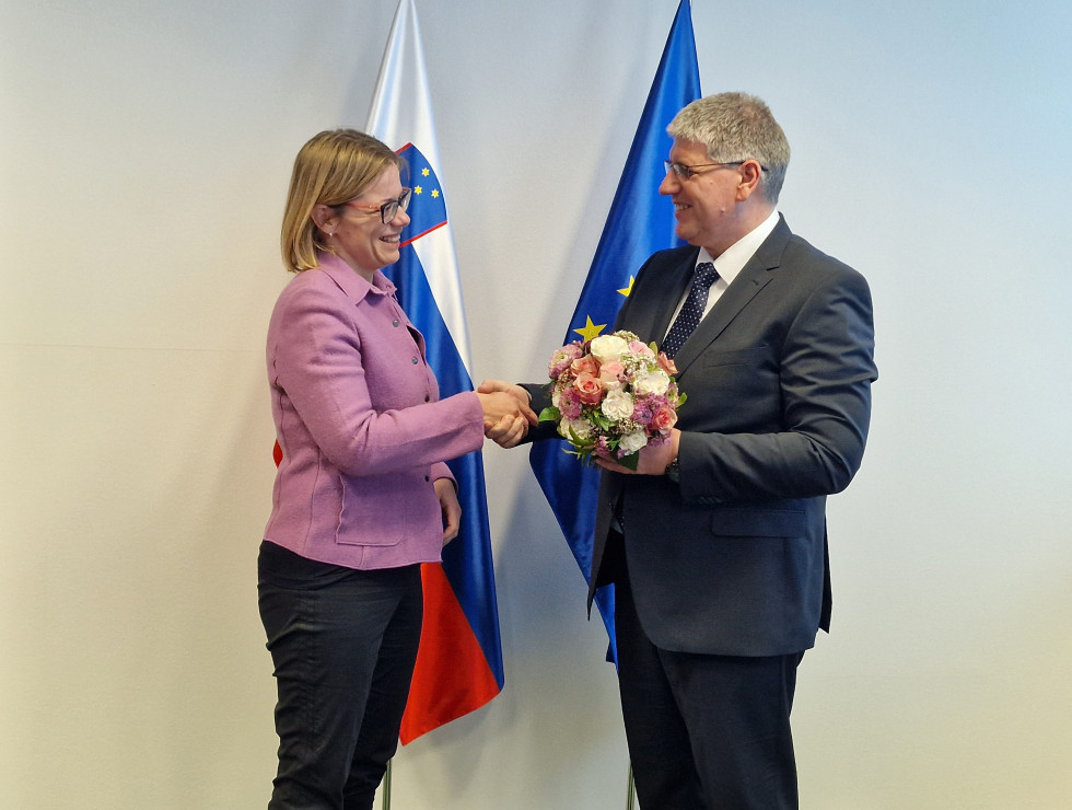 Minister Boštjan Poklukar thanked Minister Ajanović Hovnik for her work in the previous months. Giving her flowers, both standing in front of the flags.