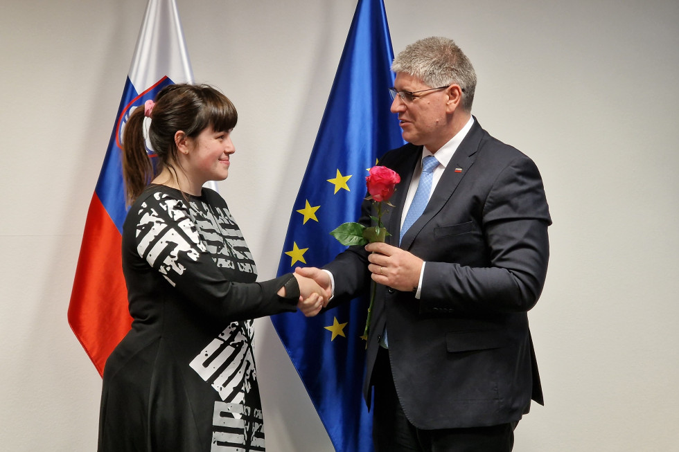 Direktorica Inštituta 8. marec Nika Kovač in minister za notranje zadeve Boštjan Poklukar stojita pred slovensko in evropsko zastavo. Se rokujeta, minister ji predaja rdečo vrtnico.