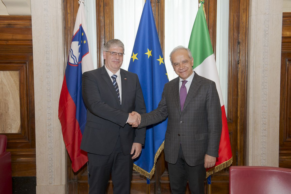 Ministra Boštjan Poklukar in Matteo Piantedosi stojita pred zastavami Slovenije, Evropske unije in Italije ter se rokujeta