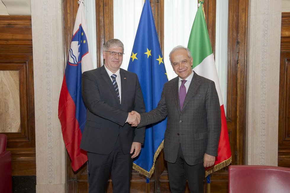 Ministers Boštjan Poklukar and Matteo Piantedosi stand in front of the flags of Slovenia, the European Union and Italy and shake hands.