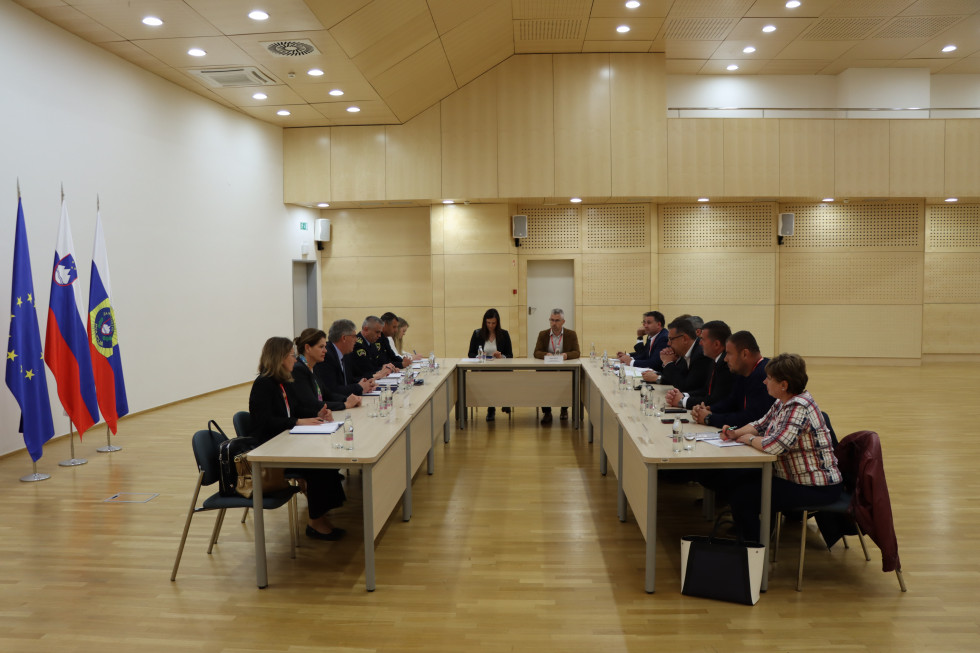 Udeleženci sestanka sedijo za mizami v obliki črke U v v dvorani. Dvorana je v svetlorjavih tonih, na stropu luči.