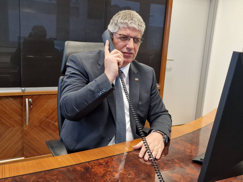 Minister Boštjan Poklukar sedi za rjavo lakirano mizo, v rokah drži telefonsko slušalko, ki jo ima prislonjeno na desno uho. Za ministrom omara, spodnja polovica je rjave barve, zgornja polovica temno steklo, poleg bela vrata.
