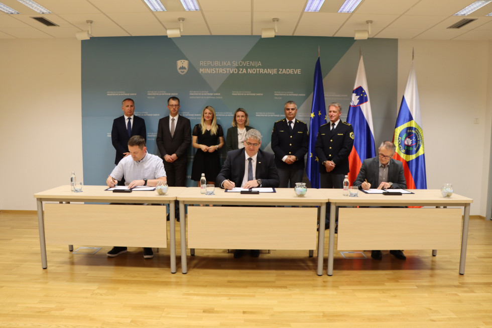Podpisniki za mizo podpisujejo sporazum, v ozadju vodstvo MNZ, zadaj evropska, slovenska in policijska zastava