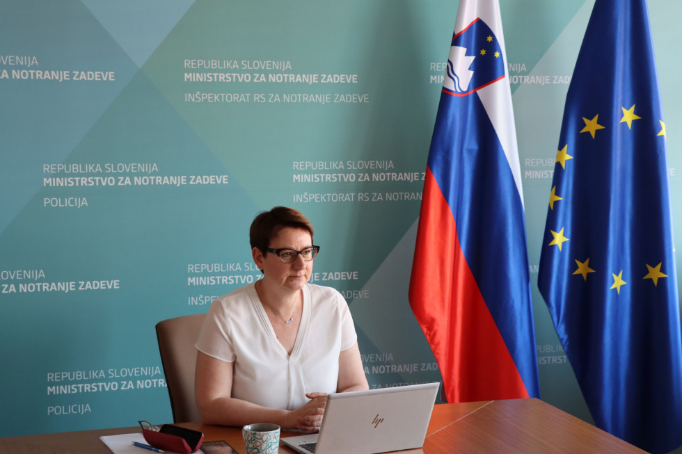 Suzana Ivanović sedi na rjavem stolu, za njo je modra stena z napisom Republika Slovenija, Ministrstvo za notranje zadeve, zadaj na njeni desni stojita slovenska in evropska zastava. Pred njo rjava miza.