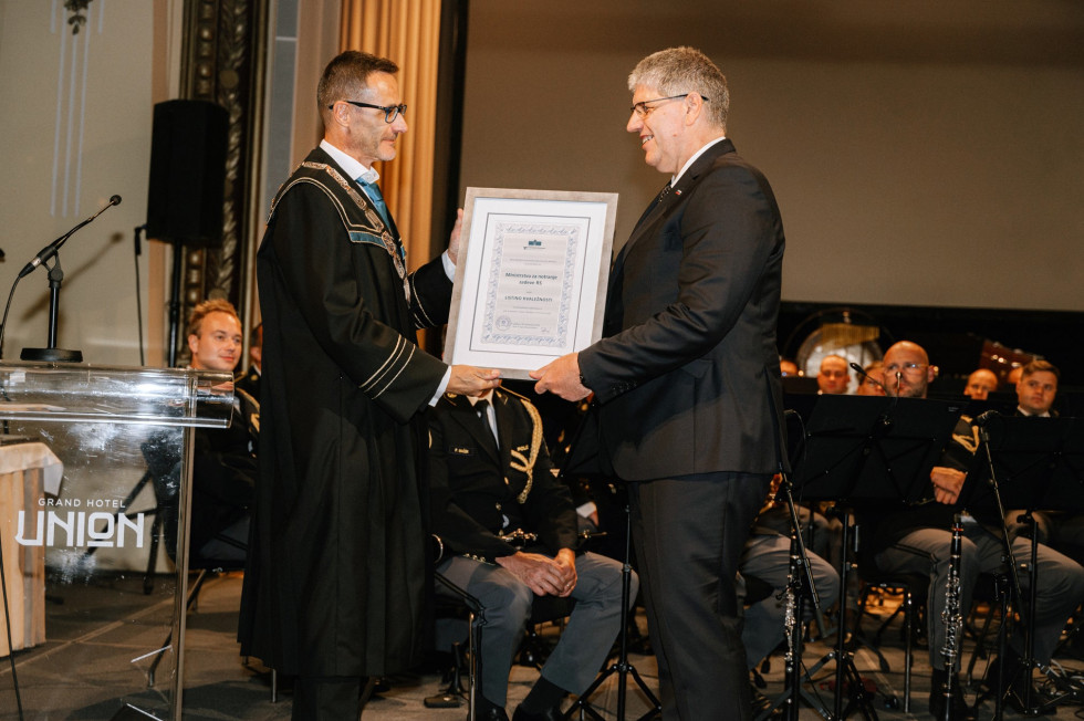 Dekan Fakultete za varnostne vede prof. dr. Igor Bernik je listino hvaležnosti slovesno podelil ministru za notranje zadeve Boštjanu Poklukarju. Dekan ministru izroča uokvirjeno listino. Oba v temnih oblekah, stojita na odru.