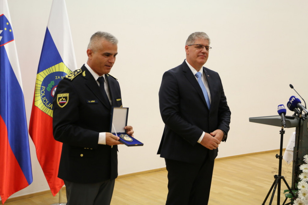 Minister Boštjan Poklukar in generalni direktor policije Senad Jušić stojita pred zastavami Slovenije, policije in Evropske unije, Senad Jušič v rokah drži znak.