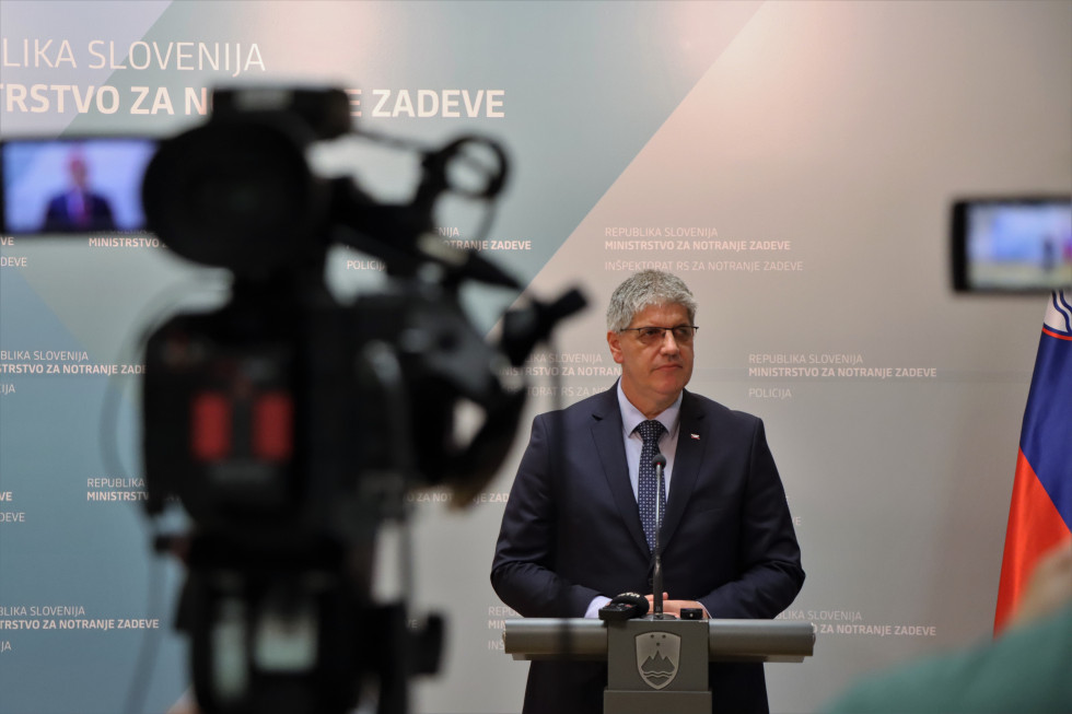 Minister of Internal Affairs Boštjan Poklukar. He stands behind a gray lectern. Behind him is a panel in blue shades. There is a recording camera in the left front corner of the photo.