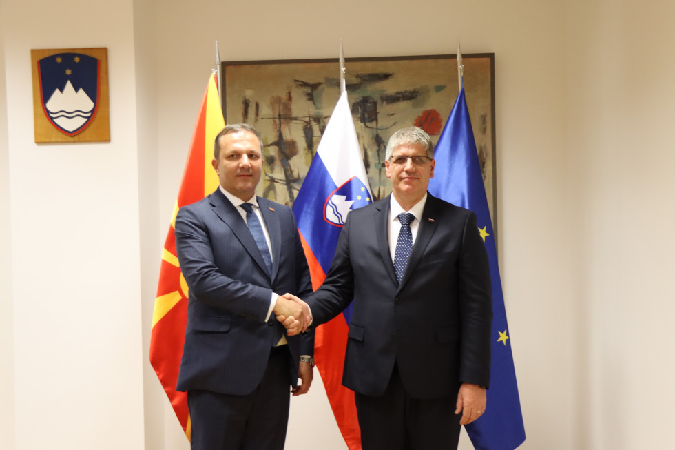 Ministers Boštjan Poklukar and Oliver Spasovski stand in front of the Macedonian, Slovenian and European flags and shake hands. 