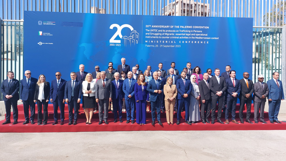 Skupinska fotografija udeležencev konference. Stojijo zunaj (približno 40 oseb), na rdeči podlagi, za njimi je moder pano z napisom 20. V ozadju modro nebo in stavbe.