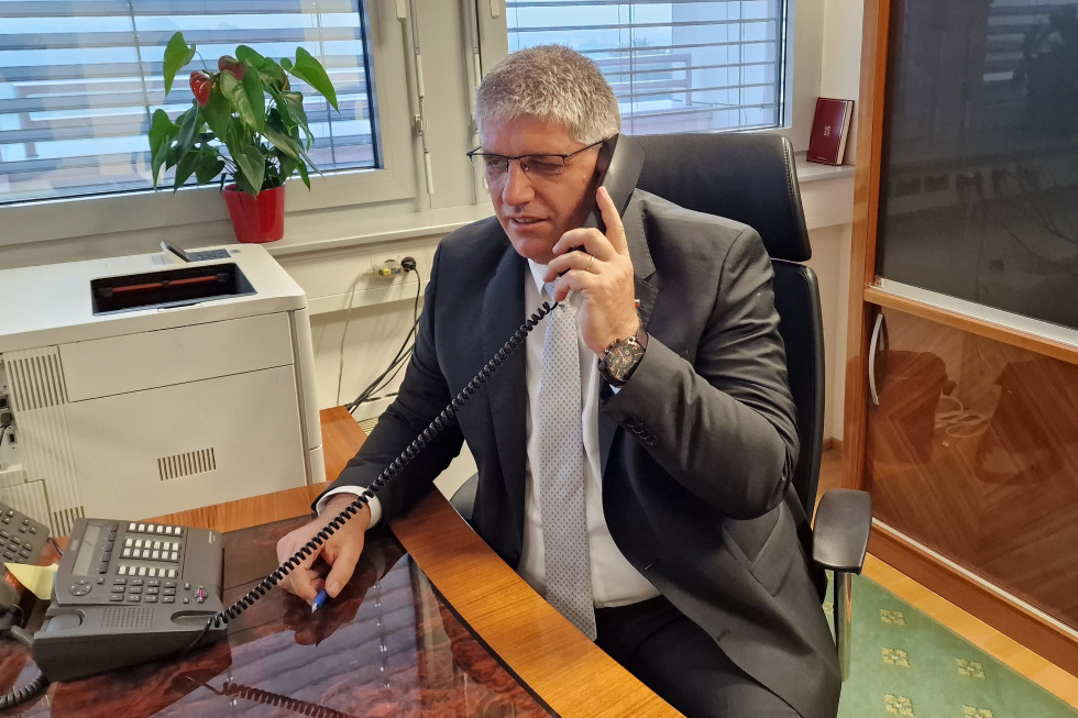 Minister za notranje zadeve Boštjan Poklukar v pogovoru s hrvaškim kolegom dr. Davorjem Božinovićem, minister sedi za mizo, v rokah drži telefonsko slušalko in govori, v ozadju na okenski polici je roža