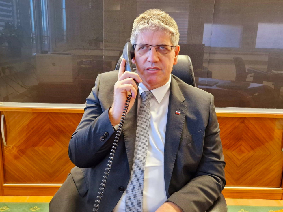 Minister of the Interior Boštjan Poklukar sits at his desk in his office. He is holding a telephone receiver. Behind him is a cabinet with a display case.