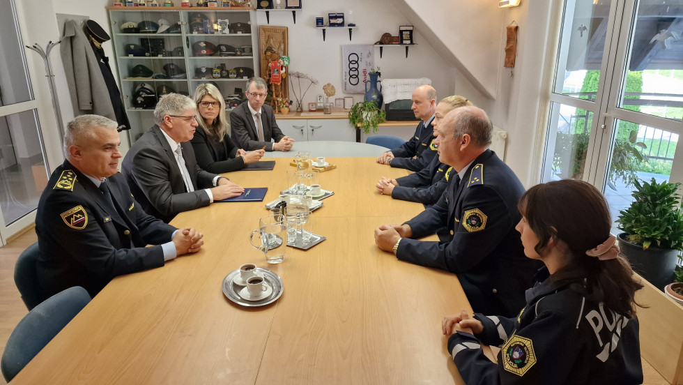 Delegacija sedi za svetlo rjavo mizo: predstavniki ministrstva na eni strani, policisti na drugi strani. Desno so okna, levo zadaj je steklena vitrina s policijskimi kapami.