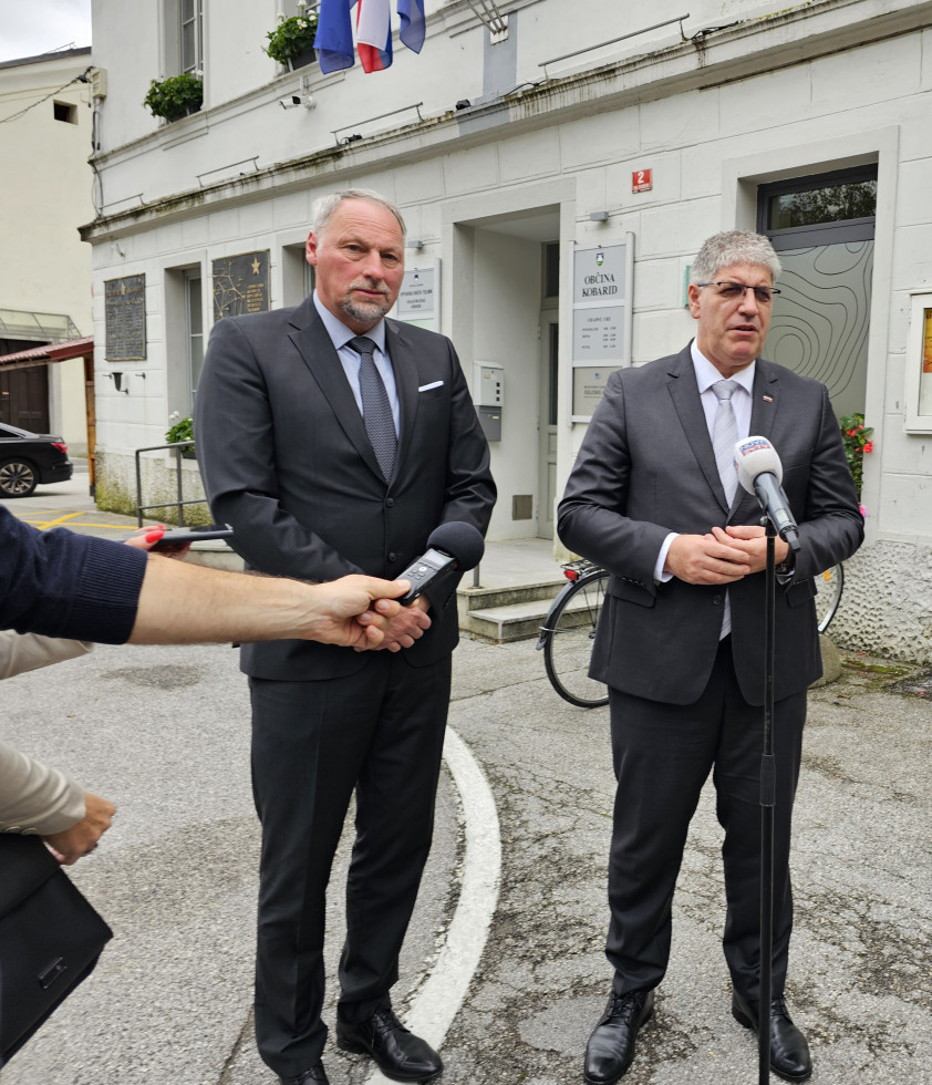 Stojita na prostem na pločniku. Pred njima stoji mikrofon na stojalu in roka, ki drži mobitel. Minister govori, župan posluša.