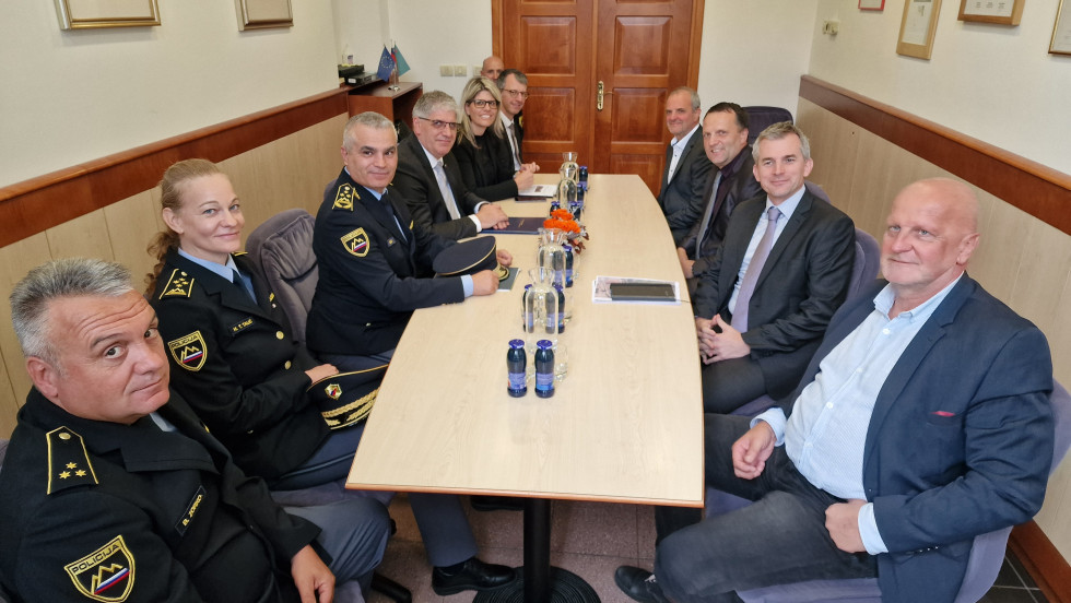 Ljudje sedijo drug ob drugem za mizo in poslušajo, na mizi so kozarci in vrči vode