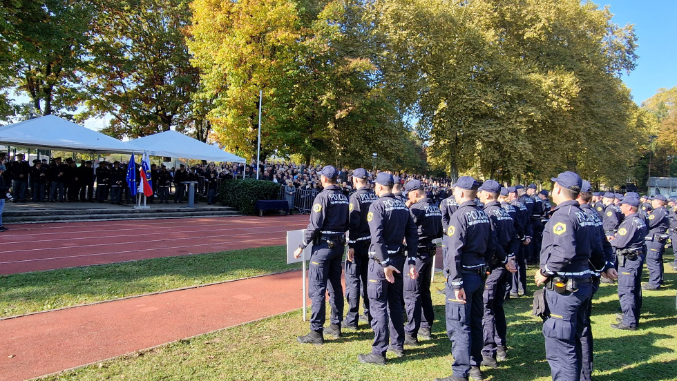 Na travi stojijo policisti drug ob drugem v vrstah, pred njimi na tleh tartan, na katerem stojijo ljudje in jih gledajo