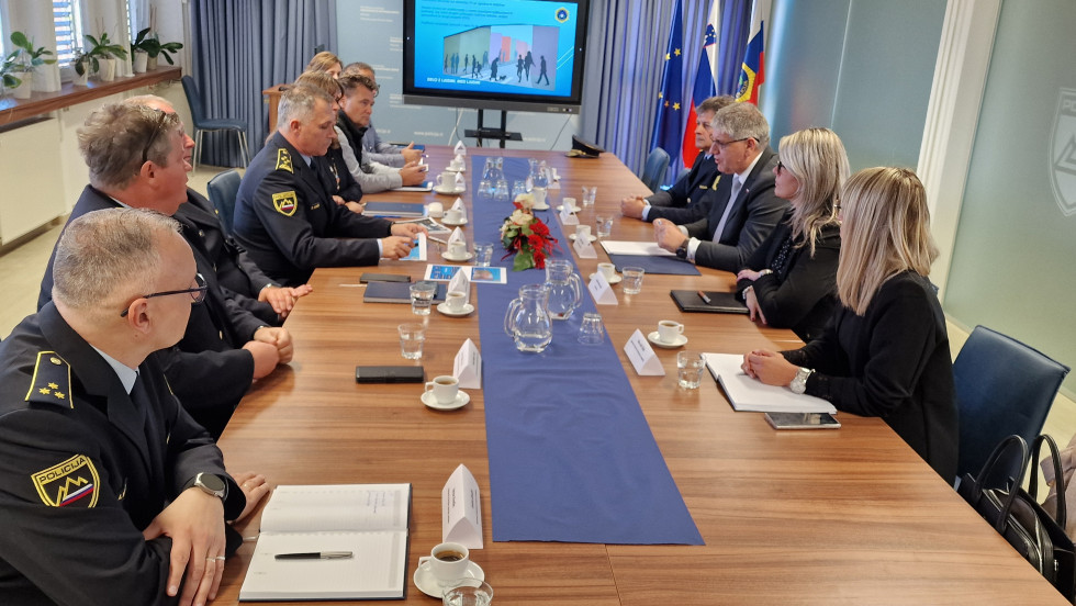 Udeleženci sedijo eden nasproti drugega za dolgo rjavo mizo. Na sredini je ozek moder prt z rožami v vazi. Na steni je projektor, poleg njega zastav.e Evropske unije, Slovenije, Ministrstva za notranje zadeve in občine Murska Sobota.
