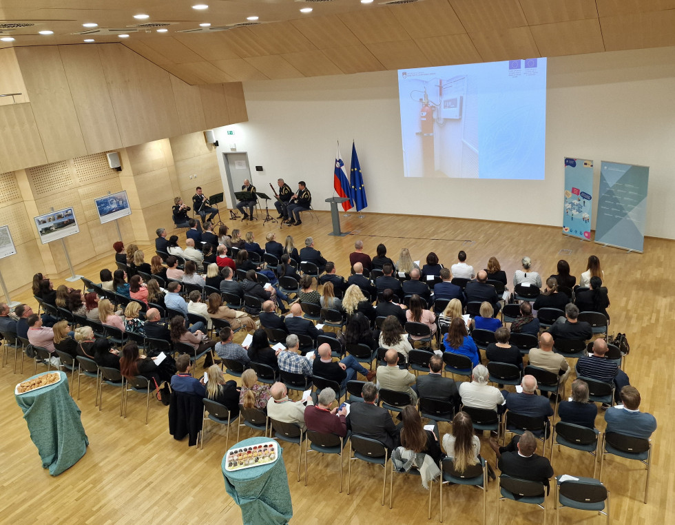 Ljudje sedijo na stolih v več vrstah in gledajo naprej, pred belo steno sedi kvintet policijskega orkestra, stojita slovenska in evropska zastava, na steni se predvajajo fotografije