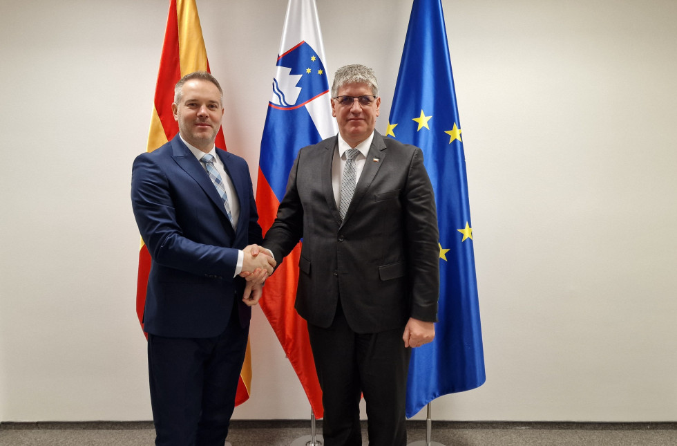 Minister in šef policije Severne Makedonije stojita pred zastavami Makedonije, Slovenije in Evropske unije in se rokujeta. Stena je bele barve.