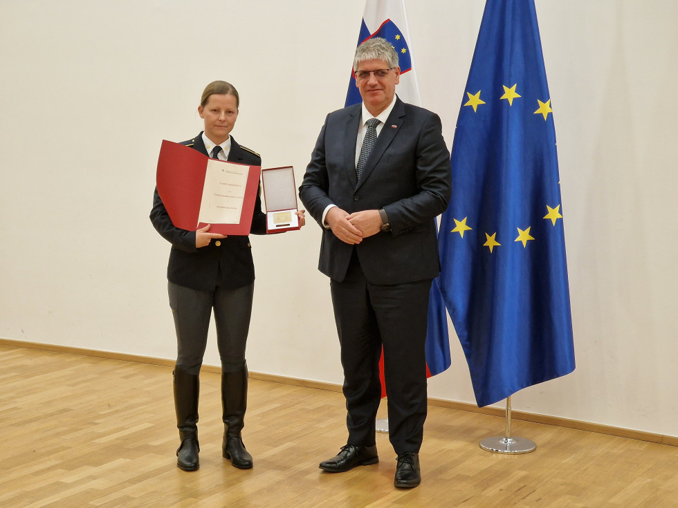 Minister in pomočnica načelnika stojita pred slovensko in evropsko zastavo, ki je ob beli steni. Pomočnica načelnika ima v roki plaketo.