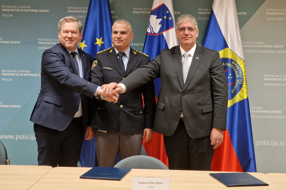 Stojijo za svetlo rjavo mizo in se rokujejo. Za njimi so tri zastave: zastava Evropske unije, slovenska in policijska. So nasmejani.