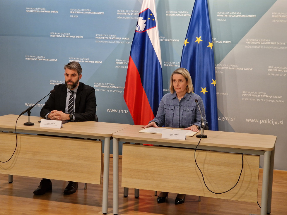 Državna sekretarka Helga Dobrin in generalni direktor Direktorata za migracije Matej Torkar sedita za svetlo rjavo mizo. Za njima je modra stena ter zastavi Slovenije in Evropske unije.