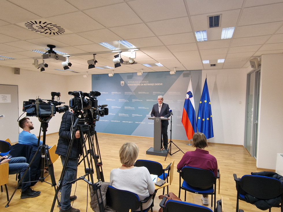 Minister za notranje zadeve Boštjan Poklukar stoji pred govornico, za njim sta slovenska in evropska zastava, v ospredju drug ob drugem sedijo novinarji in poslušajo, na levi je kamera, ki snema govorca