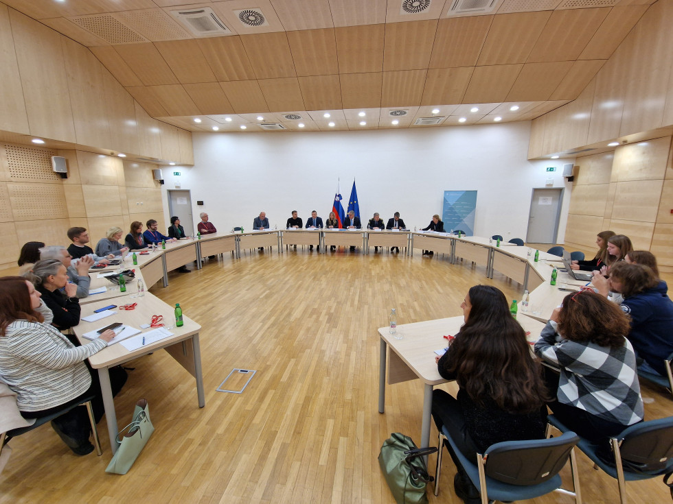 Udeleženci sedijo v dvorani za mizami v obliki črke O, na tleh je parket, mize so bež barve, zadaj ob beli steni sta slovenska in evropska zastava
