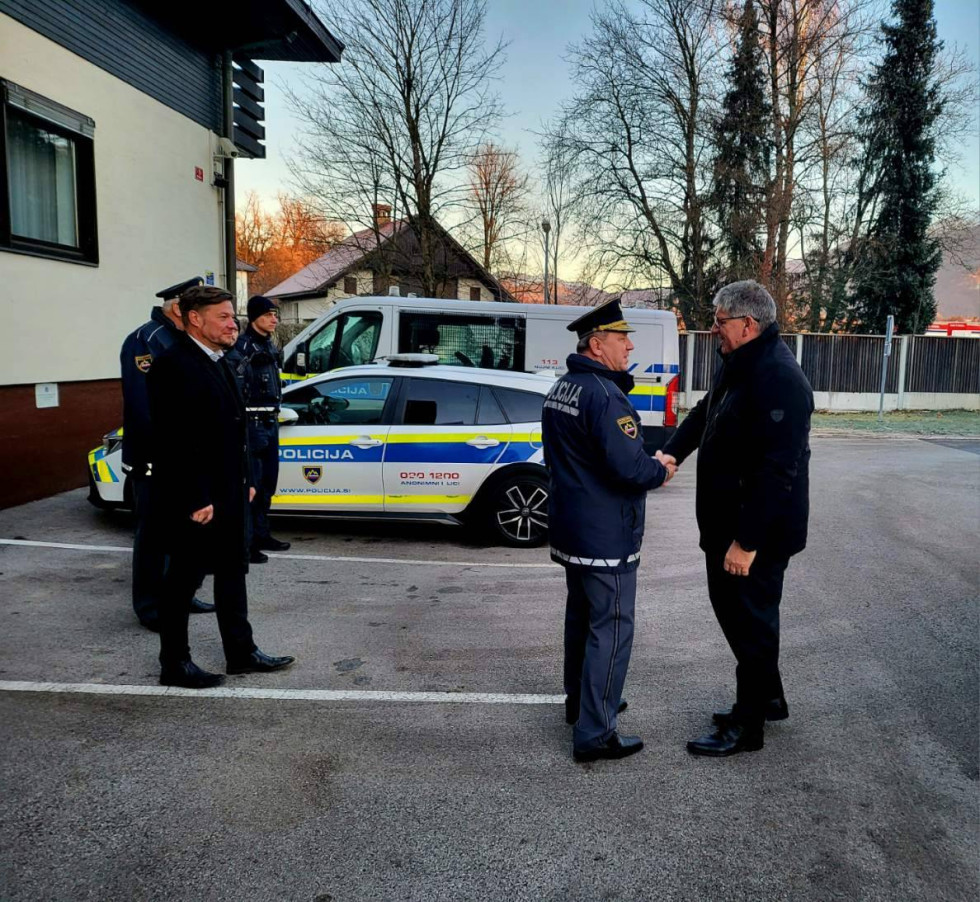 Minister se rokuje s policistom pred stavbo policijske postaje na asfaltu, v ozadju je policijski avto