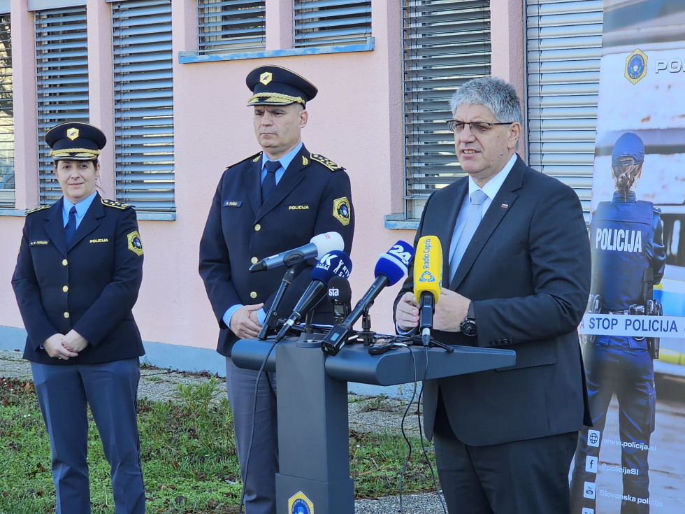 Minister stoji za govornico in govori, na govornici so postavljeni mikrofoni, ob njem stoji generalni direktor policije in policistka