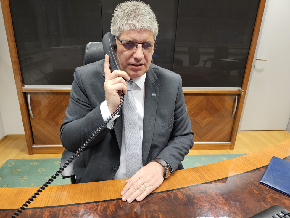 Sedi v pisarni za mizo, v roki drži telefonsko slušalko in se pogovarja po telefonu. Za njim je vitrina s temnim steklom.