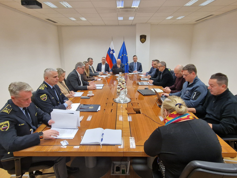 Udeleženci sedijo za podolgovato rjavo mizo drug nasproti drugega. Stene so bele barve.