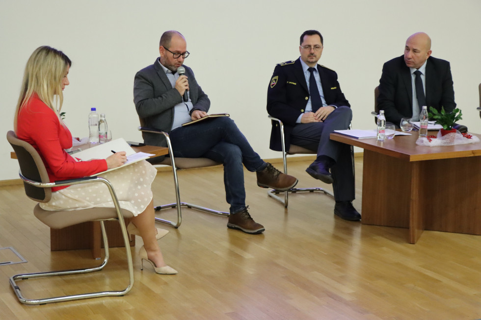 Panelisti sedijo na stolu pred belo steno, pred njimi je rjava miza.