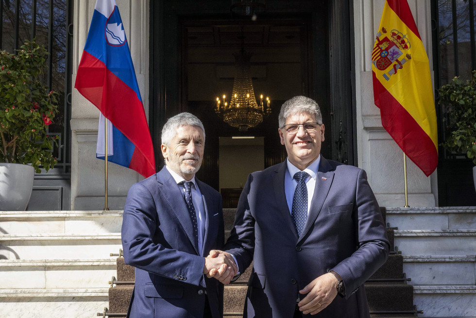 The two ministers are standing on the stairs shaking hands, with a large chandelier behind them, a Slovenian flag in the left corner and a Spanish flag in the right.